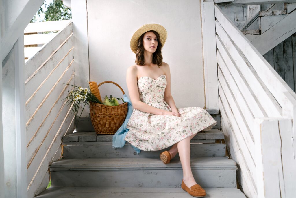 A woman in a floral dress and straw hat sits with a picnic basket on rustic wooden steps.