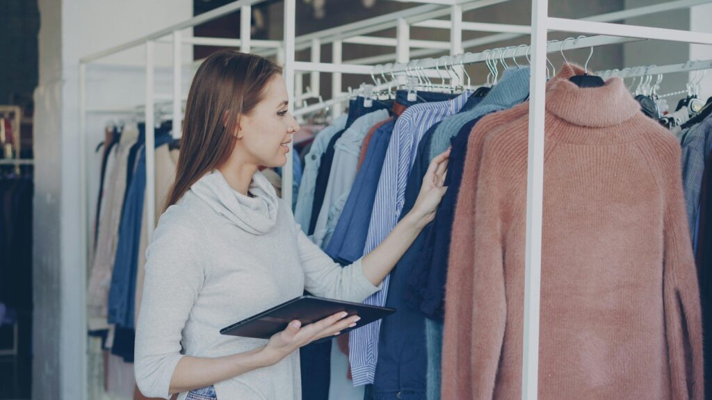 A woman browsing sweaters in a fashion retail store holding a tablet.