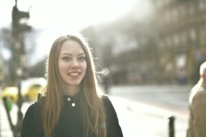A young woman smiling brightly outdoors on a sunny street.